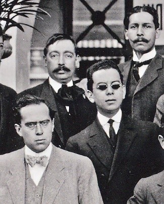 Fragmento de fotografía que muestra a dos periodistas con anteojos sin aro de tipo refinado con lentes incoloros primera fila inferior a la izquierda y con lentes obscuros