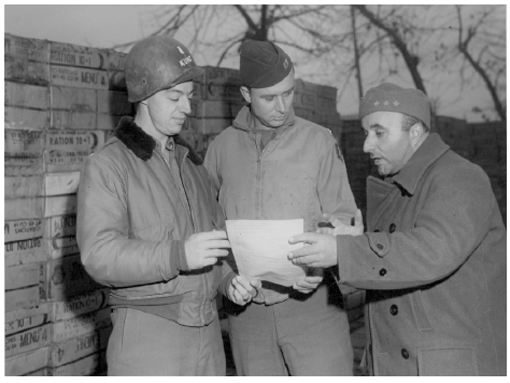 O capitão Daniel Cristóvão, do Rio de Janeiro, discute as necessidades alimentares da Força Expedicionária Brasileira (FEB) junto com os colegas norte-americanos, o tenente Kunz e o capitão Kirst, em novembro de 1944, com o inverno à espreita. Atrás deles, uma verdadeira parede de caixas contendo a ração enlatada “10 por 1”.