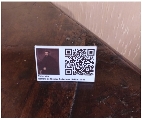 Detalhe da placa, n�o fixa, disposta em cima de um mobili�rio em exposi��o, contendo QR Code que conecta a um texto informativo amplo.