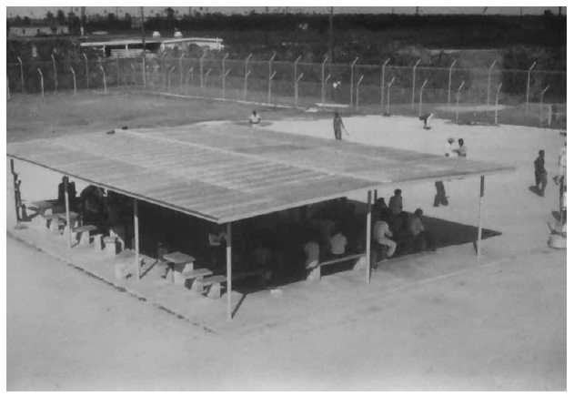 Patio de recreo en el PIDC, aprox. 1981. Debido al intenso calor en el RGV, el INS proporcionó un área de sobra para los detenidos en el patio de recreo detrás del original Centro de Detención de Port Isabel. Cerca de una docena de toldos más pequeños eventualmente quedaron desperdigados por la propiedad. Los agentes de detención vigilaban a los detenidos en el patio desde su torre de observación. PM (Fotografía 11.206f, aprox. 1981, n.d., p.206; Fotografía 11.206i, aprox . 1981, n.d.; Fotografía 11.206j, aprox. 1981, n.d.).
