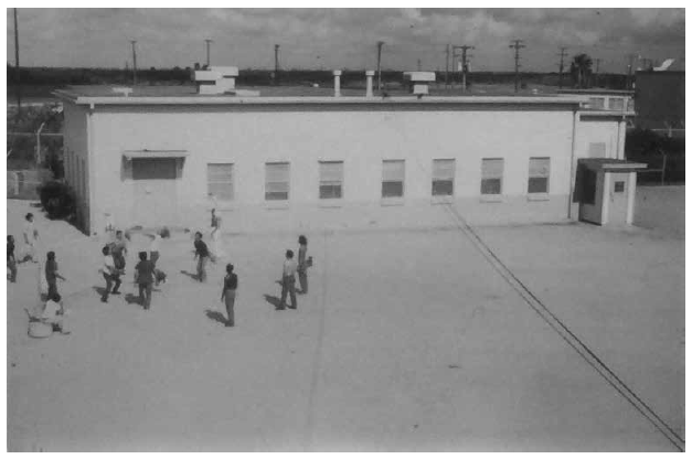 Patio de recreo en el PIDC, aprox. 1981. Debido al intenso calor en el RGV, el INS proporcionó un área de sobra para los detenidos en el patio de recreo detrás del original Centro de Detención de Port Isabel. Cerca de una docena de toldos más pequeños eventualmente quedaron desperdigados por la propiedad. Los agentes de detención vigilaban a los detenidos en el patio desde su torre de observación. PM (Fotografía 11.206f, aprox. 1981, n.d., p.206; Fotografía 11.206i, aprox . 1981, n.d.; Fotografía 11.206j, aprox. 1981, n.d.).
