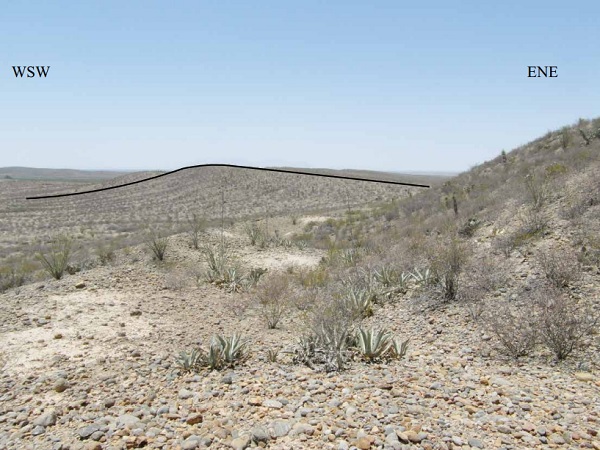 A large portion of the surface geology is hidden under a thin, but continuous cover of desert pavement, composed mainly by well-rounded clasts (pebbles to small cobbles) mainly derived from Mesozoic sedimentary rocks and a small proportion of Cenozoic(?) volcanic rocks. The foreground shows desert pavement. Near the horizon, gently inclined cuestas indicate that the succession is tilted to the ENE: the asymmetry in profile of the cuestas allow to infer the dip direction of the underlying layers in the volcano-sedimentary succession.