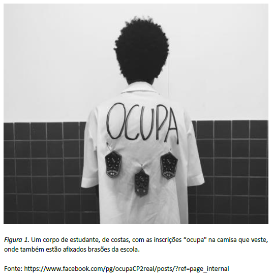 Um corpo de estudante, de costas, com as inscrições “ocupa" na camisa que veste, onde também estão afixados brasões da escola.