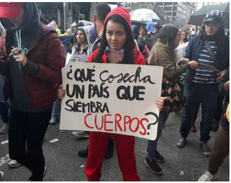 Mujer en la marcha del 21 de noviembre, en la carrera s&eacute;ptima de Bogot&aacute;, con un cartel que dice: "Qu&eacute; cosecha un pa&iacute;s que siembra cuerpos?"